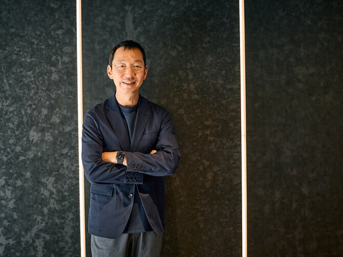 黒いテクスチャの壁を背景に、腕を組んで穏やかに微笑む山口周氏。