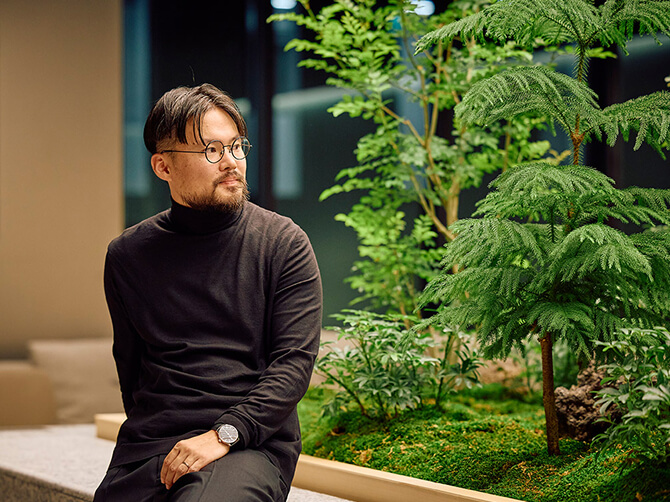オフィスラウンジの緑あふれる植栽のそばに座り、静かに遠くを見つめている湯山皓氏。