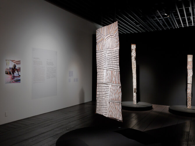 ノンギルンガ・マラウィリによるバーク・ペインティングが展示室中央に吊り下げられ、周囲には縦長の彫刻作品が並ぶ空間。