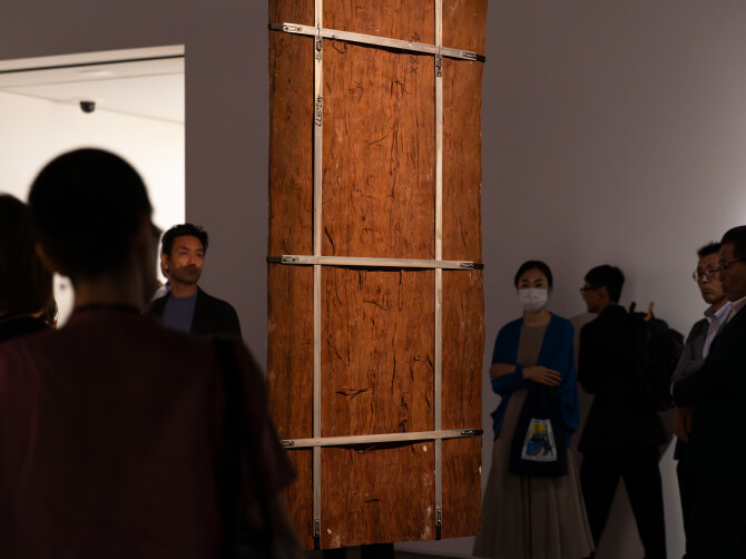 ユーカリの樹皮を素材にしたバーク・ペインティングの裏側が見える位置から、鑑賞者が作品を囲んで見つめている様子。