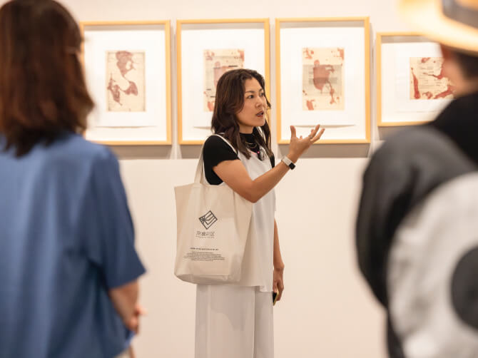 上田杏菜氏が観客に向けて作品解説を行い、背景にジュディ・ワトソンのシリーズ作品が額装されて展示されているギャラリー内の様子。