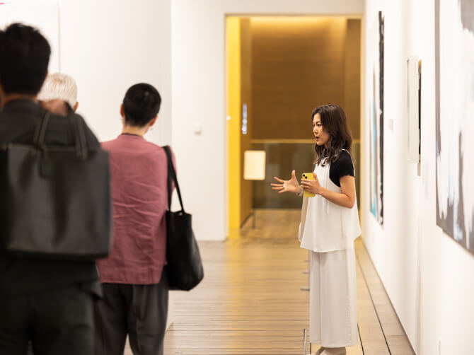 上田杏菜氏が抽象画の前で身振りを交えながら参加者に作品の背景や制作意図を説明している。