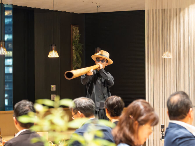 カフェテリアでデジュリドゥを演奏するGOMA氏が、客席に向けて長い木製管楽器を吹いている様子。