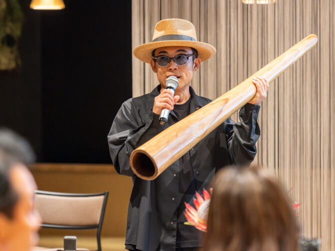 GOMA氏がマイクを手に作品や演奏について説明しながら、楽器を観客に見せている。