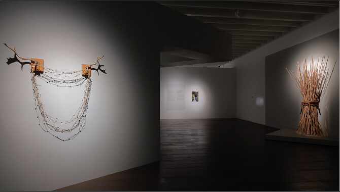 暗い展示室に、壁面に掛けられた鹿の角とチェーン状の素材を使った立体作品と、奥に木材を束ねたような彫刻作品が照明で浮かび上がって見える。