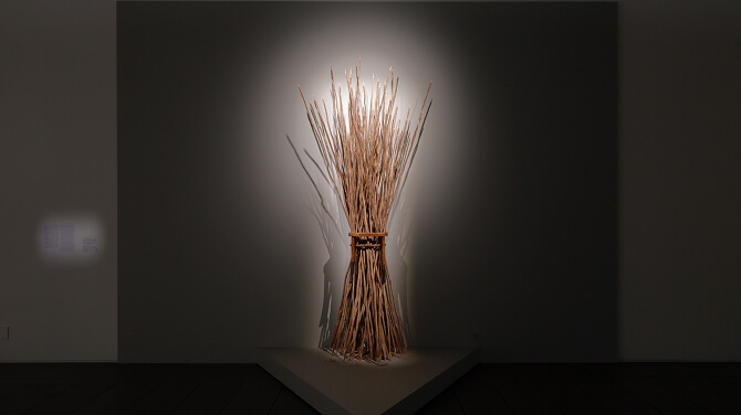 壁面に向けて照明が当てられ、細長い木材を束ねて作られた彫刻作品が立体的に強調されている。影が後ろの壁に大きく伸び、素材の質感が際立っている。