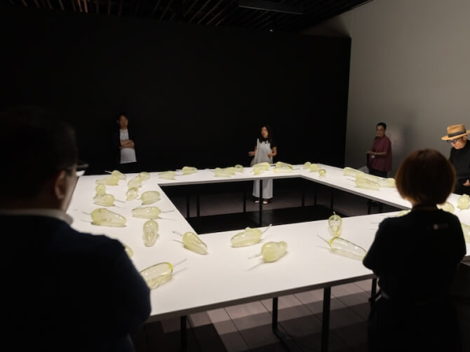 白いテーブルを囲んで観客が立ち、テーブルの上にはライトに照らされた透明なガラス作品が並んでいる。奥で上田杏菜氏が話している。