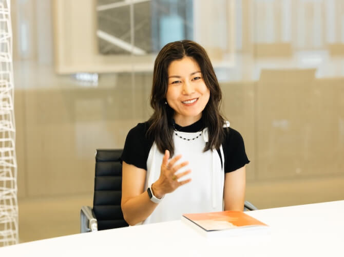 椅子に座り、笑顔で手振りを加えながら話す上田杏菜氏。テーブルの上には冊子が置かれている。