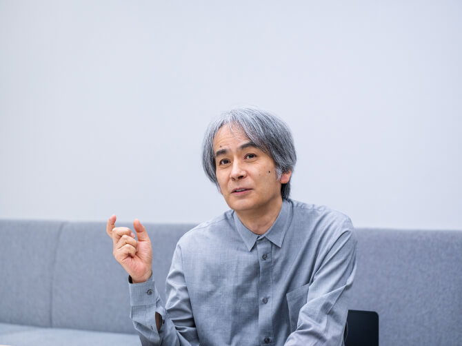 会議室のような空間で、椅子に座った高谷史郎氏が手振りを加えながら話している様子。