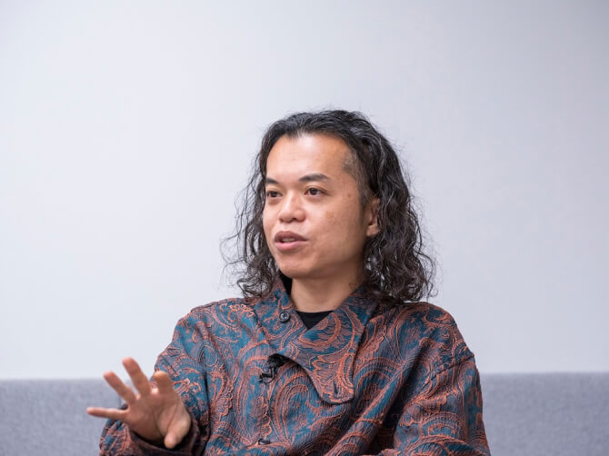 会議室のような空間で、椅子に座った古舘健氏が手振りを加えながら話している様子。