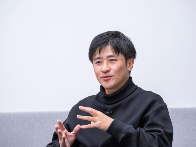 会議室のような空間で、椅子に座った濱哲史氏が手振りを加えながら話している様子。