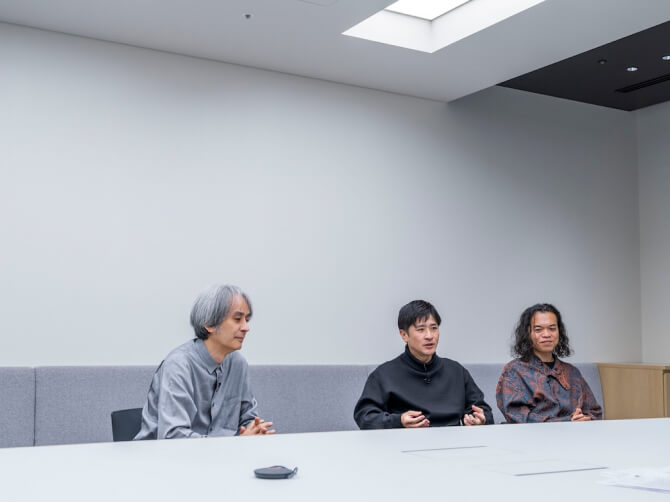 会議室のような空間で、左から高谷史郎氏、濱哲史氏、古舘健氏が並んで椅子に座り、濱哲史氏が手振りを加えながら話し、他の二人は穏やかな表情で話を聞いている様子。