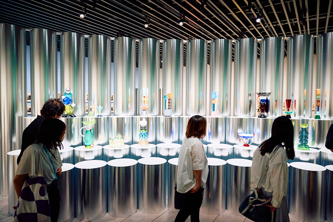 金属調の壁と台座が広がる展示室で、色鮮やかなソットサスのガラスや陶器作品を鑑賞する来場者たち。