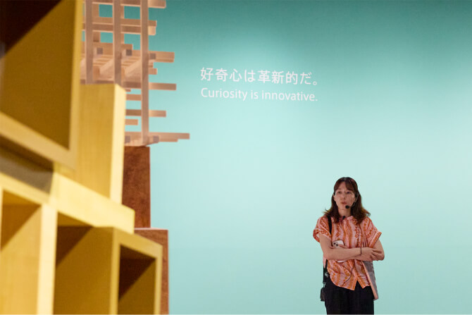 壁に掲げられたソットサスの言葉を来場者のシルエット越しに捉えた写真と、「好奇心は革新的だ。」の言葉の前で解説する杉本渚氏の写真。