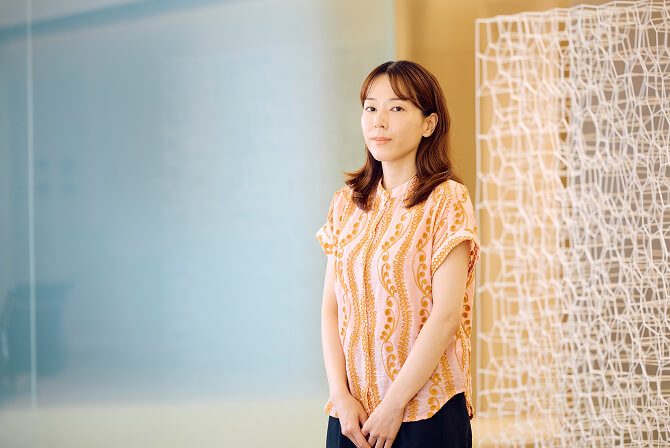 白い格子状の作品の横に立つ、オレンジ色の柄のブラウスを着た杉本渚氏のポートレート。