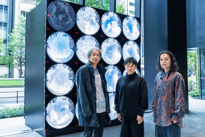 ミュージアムタワー京橋1階のエントランスに設置された《WINDOWS》の前で、左から高谷史郎氏、濱哲史氏、古舘健氏が真剣な表情で並んで立つ写真。