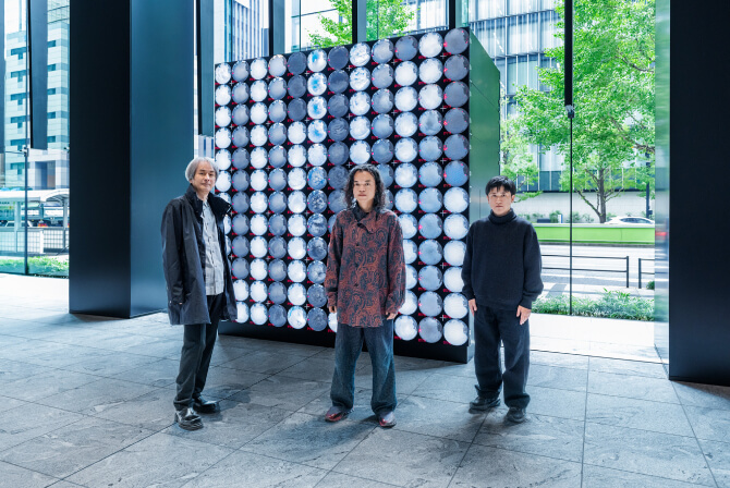 ミュージアムタワー京橋1階のエントランスに設置された《WINDOWS》の前で、左から高谷史郎氏、古舘健氏、濱哲史氏が真剣な表情で並んで立つ写真。