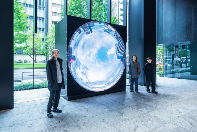 ミュージアムタワー京橋1階のエントランスに設置された《WINDOWS》の前で、左から高谷史郎氏、古舘健氏、濱哲史氏が真剣な表情で並んで立つ写真。