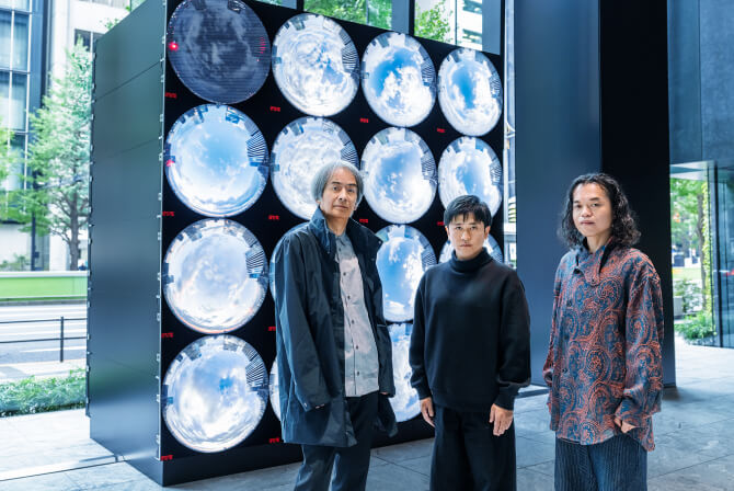 ミュージアムタワー京橋1階のエントランスに設置された《WINDOWS》の前で、左から高谷史郎氏、濱哲史氏、古舘健氏が真剣な表情で並んで立つ写真。ダムタイプのインタビュー記事「“変化するアートコレクティブ”の成り立ち」のサムネイル。