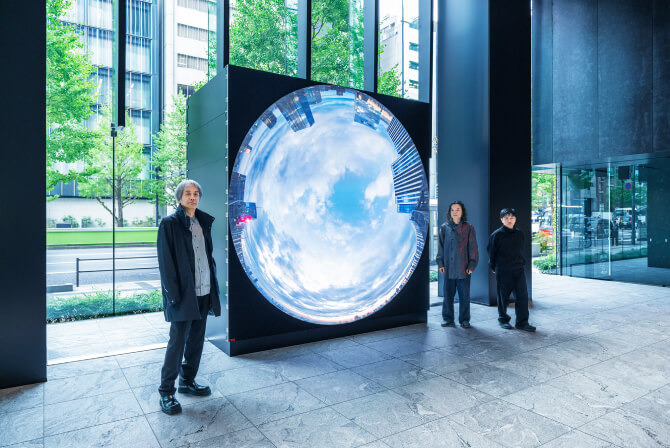 ミュージアムタワー京橋1階のエントランスに設置された《WINDOWS》の前で、左から高谷史郎氏、古舘健氏、濱哲史氏が真剣な表情で並んで立つ写真。ダムタイプのインタビュー記事「ビルそのものが世界とつながる“窓”となる」のサムネイル。