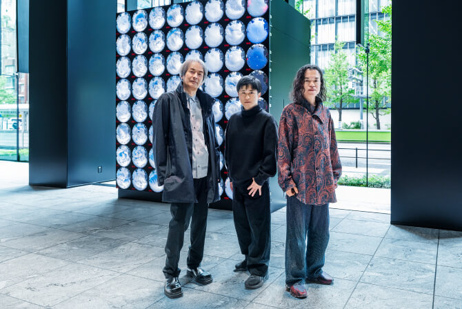 ミュージアムタワー京橋1階のエントランスに設置された《WINDOWS》の前で、左から高谷史郎氏、濱哲史氏、古舘健氏が真剣な表情で並んで立つ写真。ダムタイプのインタビュー記事「“ありのままの世界”に向き合うためのアート」のサムネイル。