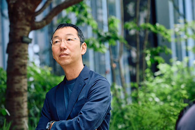 ミュージアムタワー京橋の緑の木々に囲まれた空間で、山口周氏が屋外で腕を組み、遠くを見つめている様子。山口氏のインタビュー記事「企業の一貫した美意識が社会を変える時代」のサムネイル。